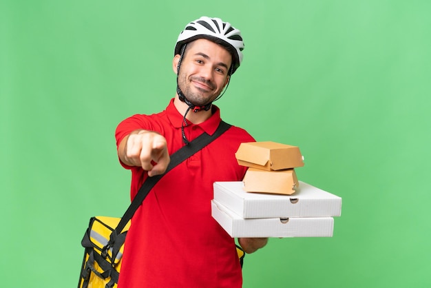 Foto joven apuesto hombre caucásico con mochila térmica y sosteniendo comida para llevar sobre un fondo aislado apuntando al frente con expresión feliz