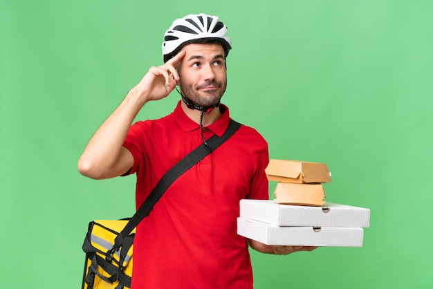 Foto joven apuesto hombre caucásico con mochila térmica y sosteniendo comida para llevar sobre un fondo aislado que tiene dudas y pensamiento