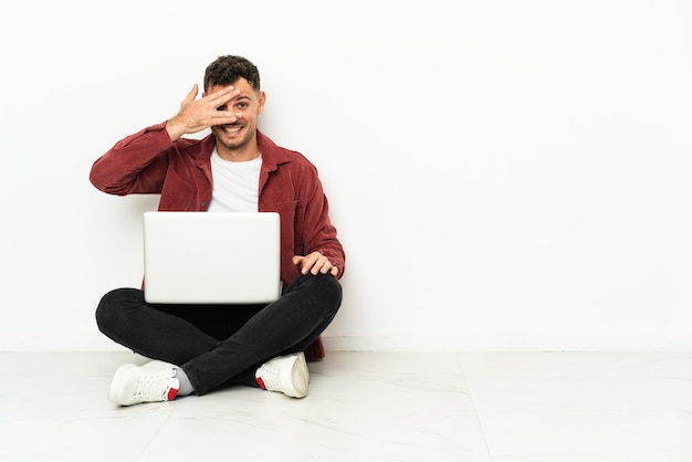 Joven apuesto hombre caucásico sentado en el suelo con la computadora portátil que cubre los ojos con las manos y sonriendo