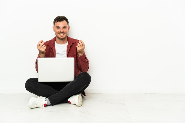 Joven apuesto hombre caucásico sentado en el suelo con laptop haciendo gesto de dinero