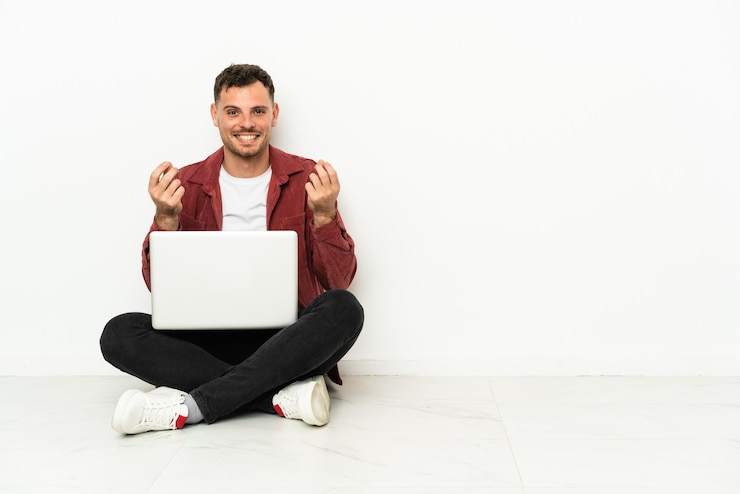 Foto joven apuesto hombre caucásico sentado en el suelo con laptop haciendo gesto de dinero