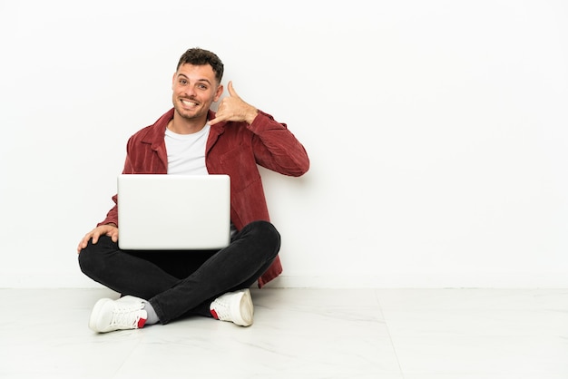 Joven apuesto hombre caucásico sentado en el suelo con laptop haciendo gesto de teléfono. Llámame señal