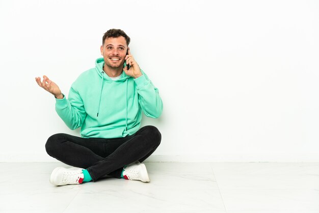 Joven apuesto hombre caucásico sentado en el suelo manteniendo una conversación con el teléfono móvil con alguien