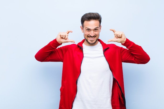 Joven apuesto hombre deportivo o monitor enmarcando o esbozando su propia sonrisa con ambas manos, mirando positivo y feliz, concepto de bienestar contra la pared plana