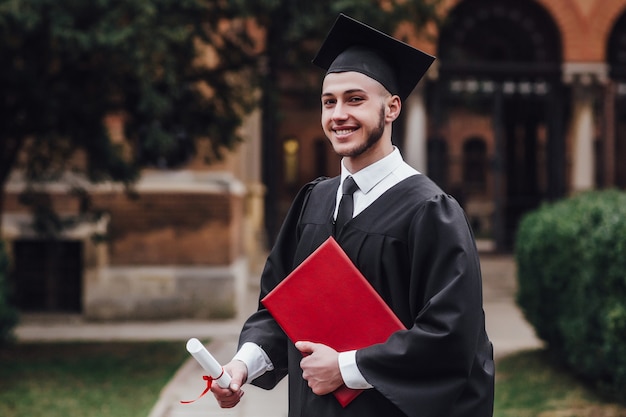 Joven apuesto hombre graduado en manto con título cerca de la universidad