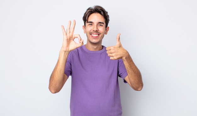 Joven apuesto hombre hispano que se siente feliz, asombrado, satisfecho y sorprendido, mostrando gestos bien y pulgares arriba, sonriendo