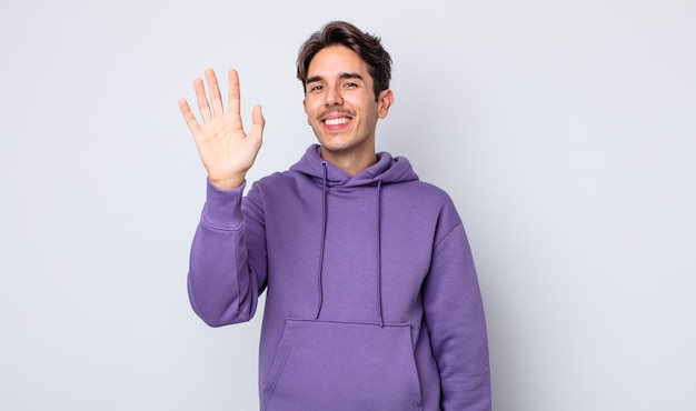 Joven apuesto hombre hispano sonriendo feliz y alegremente, saludando con la mano, dándote la bienvenida y saludándote, o despidiéndote