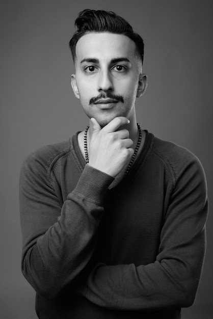 Joven apuesto hombre iraní con bigote con camisa de manga larga contra la pared gris en blanco y negro