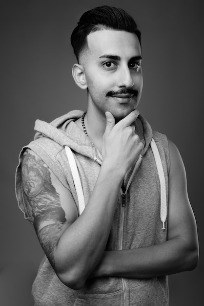 Joven apuesto hombre iraní con bigote vistiendo camisa con capucha contra la pared gris en blanco y negro