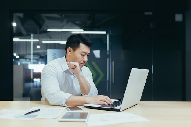 Un joven apuesto, un hombre de negocios asiático, está trabajando con una computadora portátil escribiendo sentado en un escritorio en un