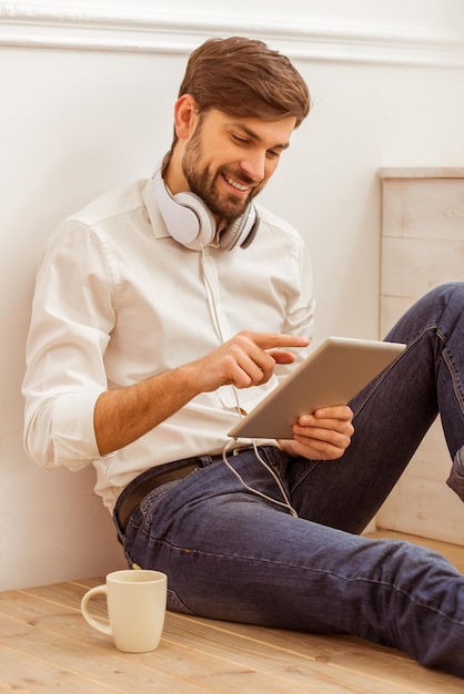 Joven apuesto hombre de negocios con camisa clásica blanca y jeans usando una tableta y sonriendo mientras se sienta en el piso de madera