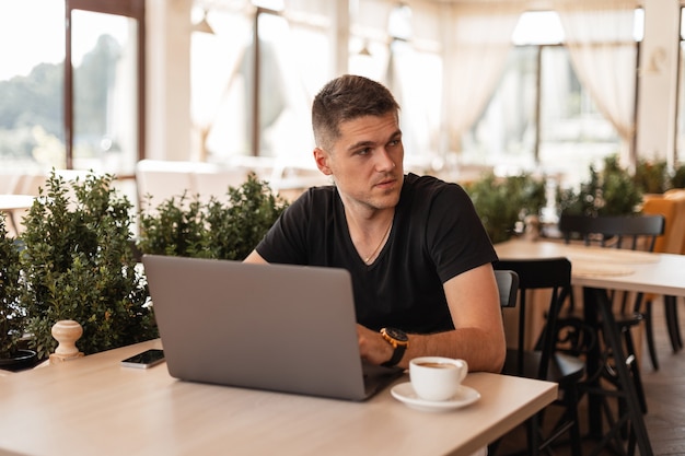 Joven apuesto hombre de negocios con una elegante camiseta negra sentado en un café y trabajando de forma remota en una computadora portátil moderna