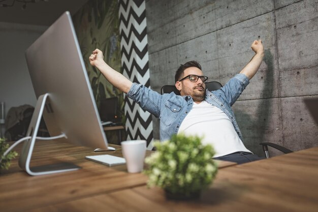 Joven apuesto hombre de negocios con exceso de trabajo sentado en la oficina estirando y esperando el final de las horas de trabajo.