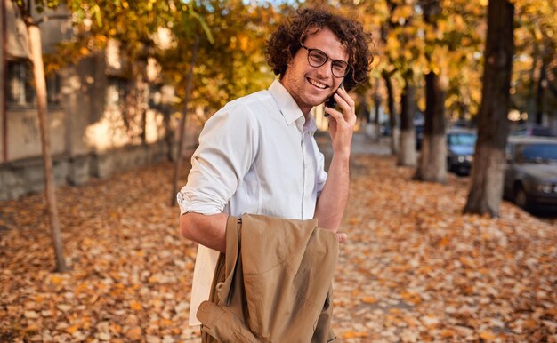 Joven apuesto hombre de negocios posando en la calle de otoño mientras camina al aire libre y usa un teléfono inteligente para llamar Hombre sonriente con cabello rizado con gafas hablando en su teléfono móvil con amigos