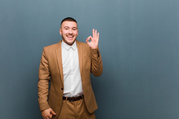 Foto joven apuesto hombre de negocios que se siente exitoso y satisfecho, sonriendo con la boca abierta, haciendo un signo de bien con la mano contra la pared plana