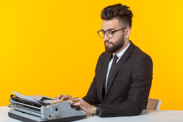 Joven apuesto hombre de negocios en ropa formal escribiendo texto en una máquina de escribir posando en una pared amarilla