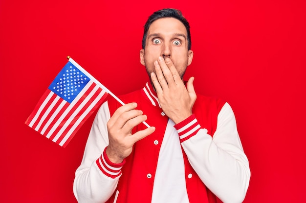 Joven apuesto hombre patriótico sosteniendo la bandera de los Estados Unidos celebrando el día de la independencia cubriendo la boca con la mano conmocionado y asustado por el error Expresión sorprendida