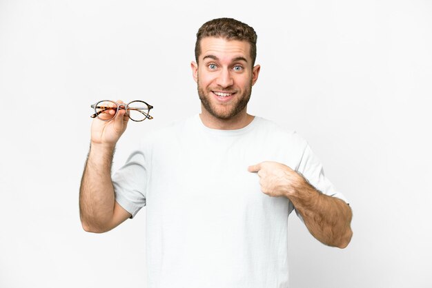 Joven apuesto hombre rubio con gafas sobre fondo blanco aislado con expresión facial sorpresa