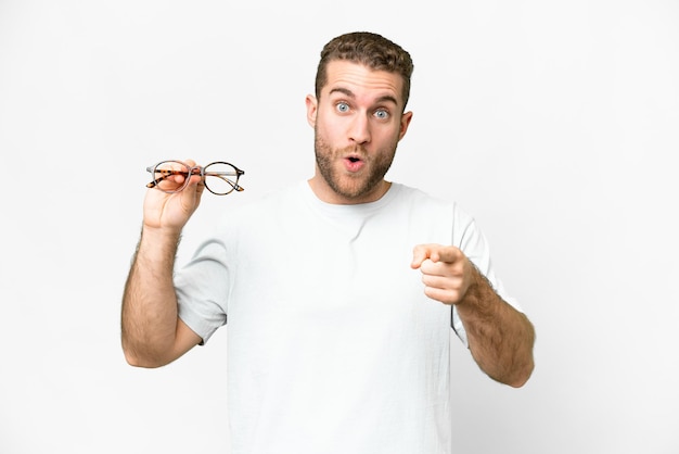 Joven apuesto hombre rubio con gafas sobre fondo blanco aislado sorprendido y apuntando al frente