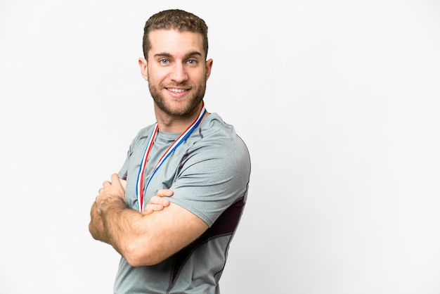 Joven apuesto hombre rubio con medallas sobre fondo blanco aislado con los brazos cruzados y mirando hacia adelante