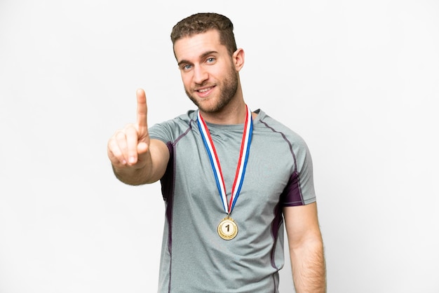 Joven apuesto hombre rubio con medallas sobre fondo blanco aislado mostrando y levantando un dedo