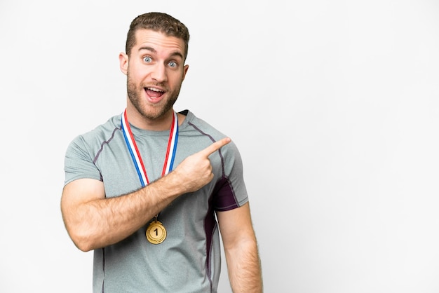 Joven apuesto hombre rubio con medallas sobre fondo blanco aislado sorprendido y apuntando hacia el lado