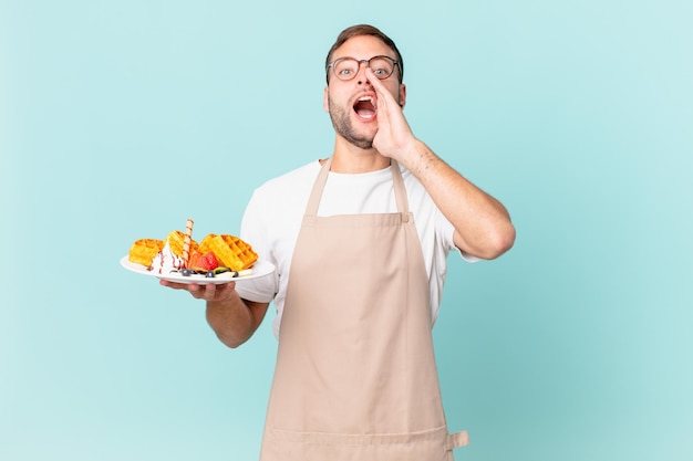 Joven apuesto hombre rubio que se siente feliz dando un gran grito con las manos al lado de la boca cocinando gofres concepto