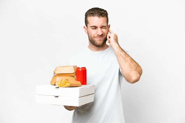 Joven apuesto hombre rubio sosteniendo pizzas y hamburguesas sobre un fondo aislado frustrado y cubriendo las orejas