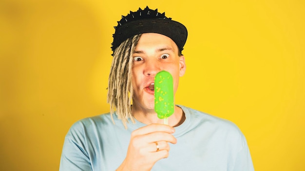 Joven apuesto hombre satisfecho con rastas en gorra negra comiendo helado sobre fondo amarillo Chico alegre en camiseta azul disfrutando de helado