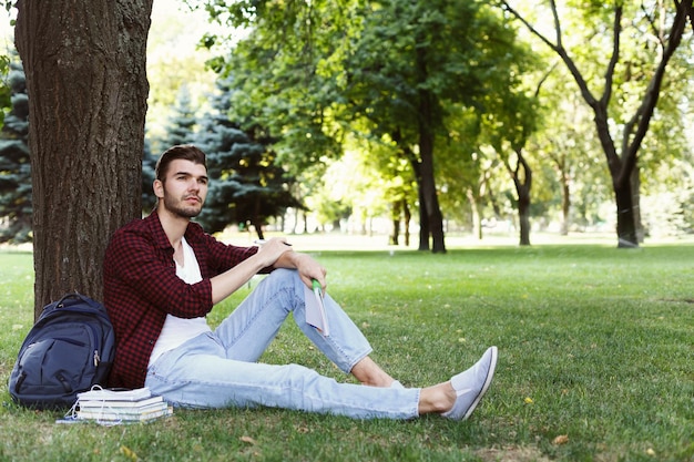 Joven apuesto hombre sonriente sentado en la hierba, descansando con libros cerca, soñando con vacaciones, copiando espacio