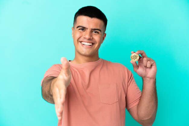 Foto joven apuesto hombre sosteniendo un bitcoin sobre fondo azul aislado un apretón de manos para cerrar un buen trato