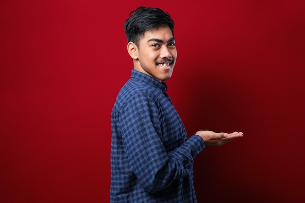 Joven apuesto hombre vestido con camisa casual sobre fondo rojo aislado apuntando a un lado con las manos abiertas palmas mostrando copia espacio, presentando publicidad sonriendo emocionado feliz