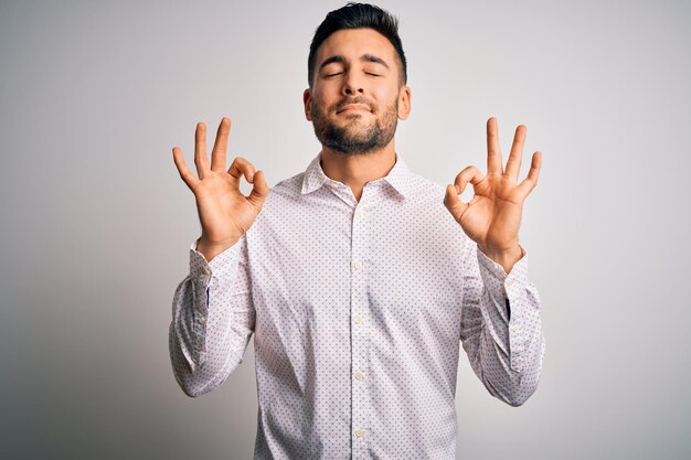 Joven apuesto hombre vestido con camisa elegante de pie sobre fondo blanco aislado relajado y sonriendo con los ojos cerrados haciendo gesto de meditación con los dedos concepto de yoga