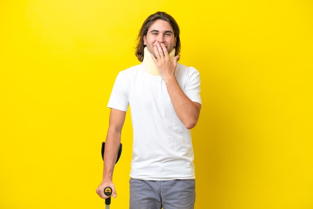 Joven apuesto hombre vestido con cuello ortopédico y muletas aislado sobre fondo amarillo feliz y sonriente cubriendo la boca con la mano