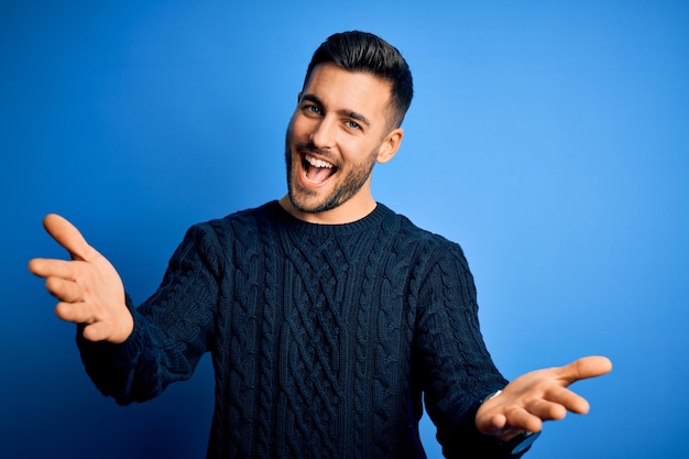 Joven apuesto hombre vestido con suéter casual de pie sobre un fondo azul aislado sonriendo alegre ofreciendo manos dando asistencia y aceptación