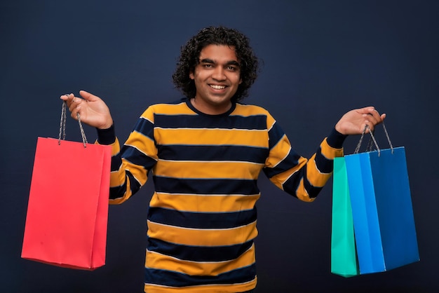 Un joven apuesto indio sosteniendo y posando con bolsas de compras en un fondo gris