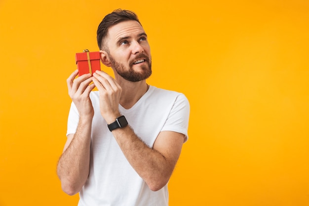 Un joven apuesto joven feliz posando aislado sobre una pared amarilla sosteniendo una caja de regalo