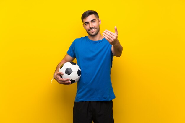 Joven apuesto jugador de fútbol hombre sobre pared amarilla aislada invitando a venir con la mano, feliz de que hayas venido