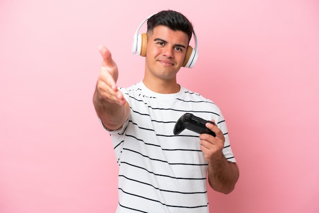 Un joven apuesto jugando con un controlador de videojuegos aislado en un fondo rosa dándose la mano para cerrar un buen trato
