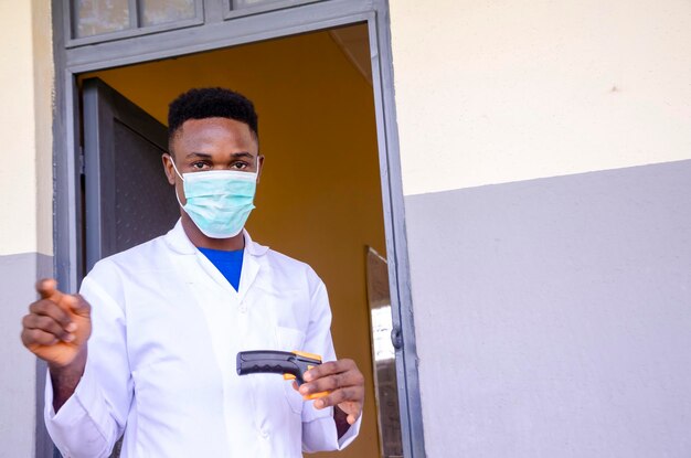 Un joven y apuesto maestro de clase africano sosteniendo como termómetro para escanear la temperatura de su estudiante antes de entrar al salón de clases