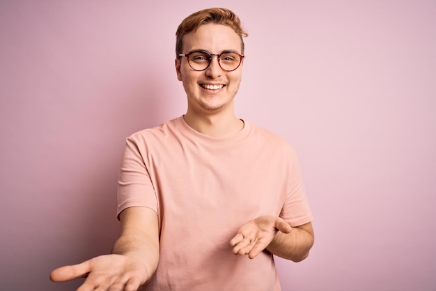 Joven apuesto pelirrojo con camiseta informal sobre un aislado fondo rosa sonriendo alegre con los brazos abiertos como bienvenida amistosa saludos positivos y confiados