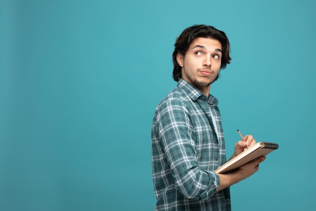 Foto un joven apuesto y pensativo parado en la vista de perfil escribiendo en un bloc de notas con un lápiz mirando a un lado aislado en un fondo azul con espacio para copiar