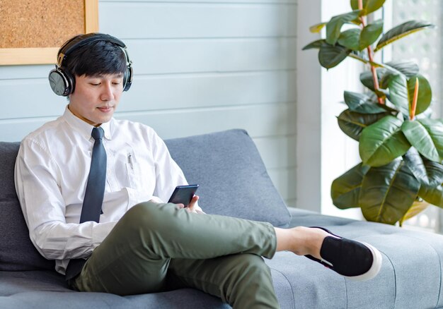 Joven y apuesto profesional asiático exitoso hombre de negocios empleado en camisa de negocios formal y corbata sentado en un cómodo sofá escuchando música en línea navegando por Internet con un teléfono inteligente.