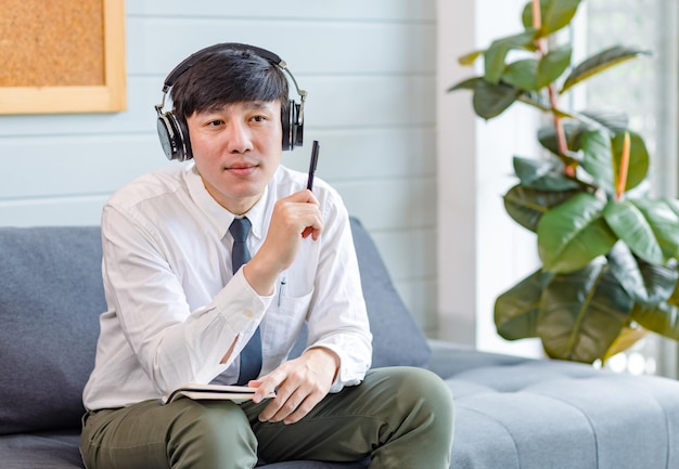 Joven y apuesto profesional asiático exitoso hombre de negocios empleado en camisa de negocios formal y corbata sentado en un cómodo sofá escuchando música en línea navegando por Internet con un teléfono inteligente.