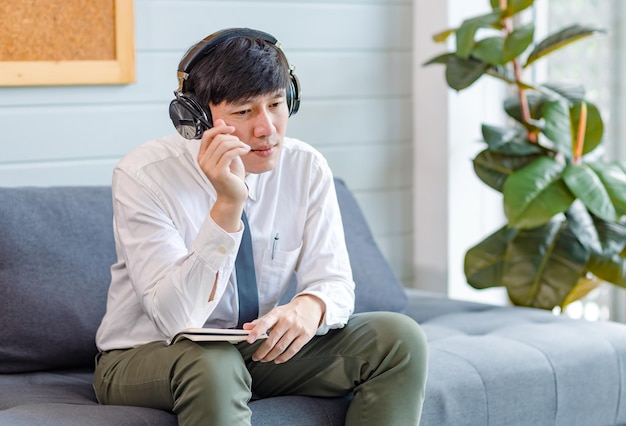 Joven y apuesto profesional asiático exitoso hombre de negocios empleado en camisa de negocios formal y corbata sentado en un cómodo sofá escuchando música en línea navegando por Internet con un teléfono inteligente.
