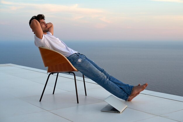 un joven apuesto que se relaja y trabaja en una computadora portátil en el balcón de su casa mientras mira la puesta de sol