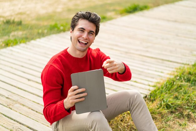 Un joven apuesto que sostiene una tableta al aire libre te señala con una expresión de confianza