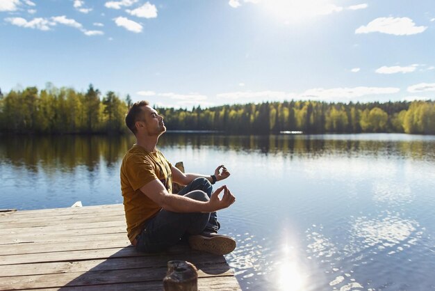 Un joven apuesto sentado cerca del lago del bosque relajándose y meditando en un día soleado de primavera o verano