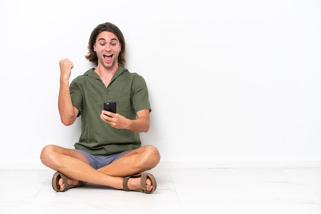 Foto un joven apuesto sentado en el suelo aislado de fondo blanco sorprendido y enviando un mensaje