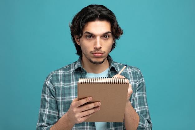 un joven apuesto serio sosteniendo un bloc de notas y un lápiz mirando a la cámara aislada de fondo azul